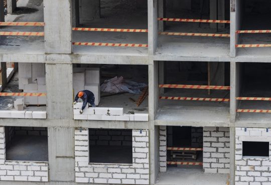 Blok mieszkalny w trakcie budowy z widocznym robotnikiem stawiającym murowaną ścianę w ramowej konstrukcji