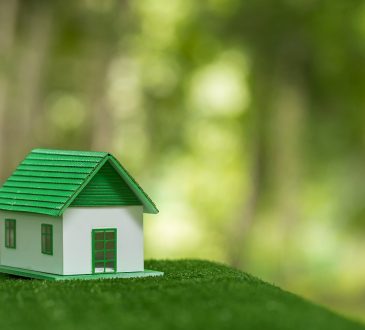 Figurka domu stojąca na trawie. W tle las rozmyty efektem bokeh